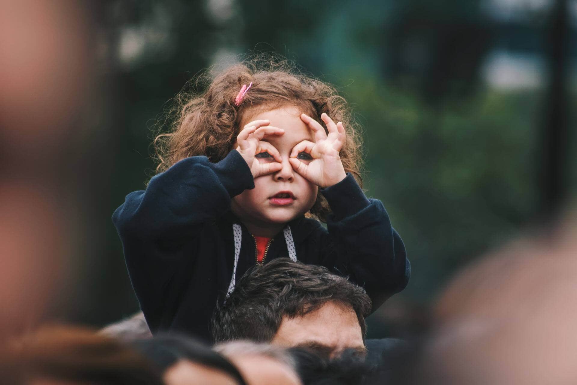 Child sitting on someone's shoulders, making binoculars with hands over eyes, outdoors in a crowd